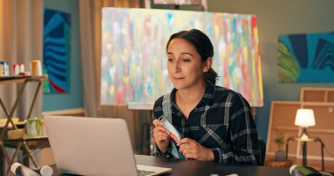 Portrait of art store employee, doing through computer, laptop live with store customers, woman showing new watercolor paints available for sale, advertising product, in background painting on easel