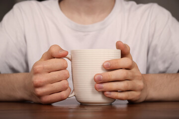 A young man sits at a table holding a hot drink in a mug