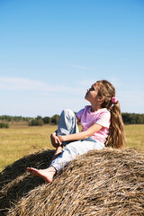 Naklejka na ściany i meble Girl sitting on haystack. Summertime in the country. Cute child on the field. Happy childhood concept.