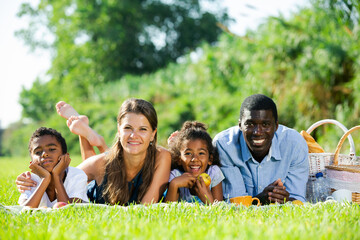 Portrait of happy interracial family with two children lying on blanket on green meadow, enjoying picnic ..