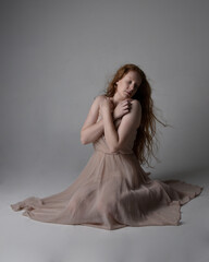 Fototapeta premium Full length portrait of pretty red haired woman dancer, wearing skin toned flowing fairy dress. Sitting gestural poses isolated on studio background.