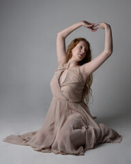 Full length portrait of pretty red haired woman dancer,  wearing skin toned flowing fairy dress. Sitting gestural poses  isolated on studio background.