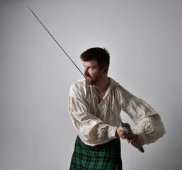 Close up portrait of handsome brunette man wearing Scottish kilt and renaissance white  pirate blouse shirt. Holding a sword weapon, action pose isolated against studio background.   