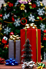 Studio shot of red and gray tall gift boxes with shiny golden ribbon placed on wooden table with colorful glossy bow tie in front decorative beautiful Christmas eve pine tree in blurred background