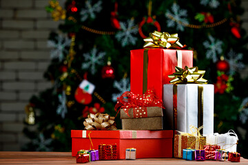 Colorful small and big present gift boxes with shiny ribbon bow tie placed on corner of wooden table in front fully decorative beautiful Christmas eve pine tree and brick wall in blurred background