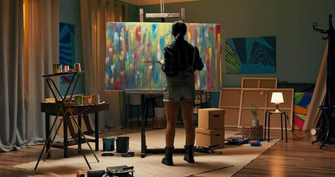 A talented artist works on an abstract painting, with long brush strokes she creates a masterpiece. A dark and untidy studio where a large canvas stands on an easel, paints on the floor and a table