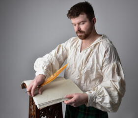 Close up portrait of handsome brunette man wearing Scottish kilt and renaissance white  pirate blouse shirt. Holding a quill and writing in a book,  pose isolated against studio background.    - obrazy, fototapety, plakaty
