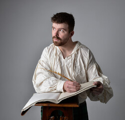 Close up portrait of handsome brunette man wearing Scottish kilt and renaissance white  pirate blouse shirt. Holding a quill and writing in a book,  pose isolated against studio background.   
