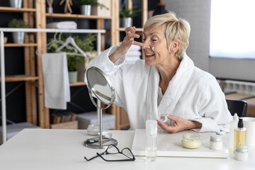 An older woman with short blonde hair in a white bathrobe sits at the table and puts cream on her face inside the living room