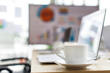 White hot tasty coffee cup on small dish placed on working table with laptop computer in meeting room in front target growth rate graph and chart report on big screen monitor in blurred background