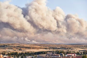 Photograph of a large forest fire with a large column of smoke on a hillside near a city