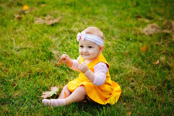 Portrait of a one-year-old smiling girl in a yellow dress sitting barefoot on a lawn against a background of green grass.