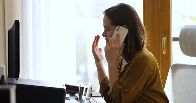 Side view young business woman sit at workplace desk lead formal conversation on phone, female looks at pc screen discuss online purchase with agent customer services. Workflow, modern tech concept
