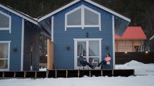 Caucasian Male and Female Chatting while Sitting on a Terrace of Scandinavian Style House. Travel, Tourism, Winter Holidays Concept