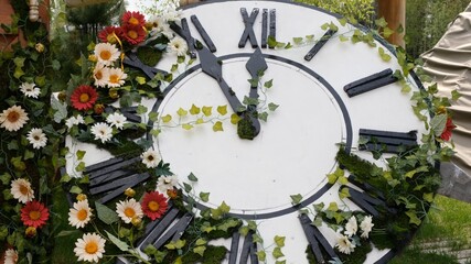 Large wall clock on the field in blooming flowers, showing the time 11:55, 12:00. time management...