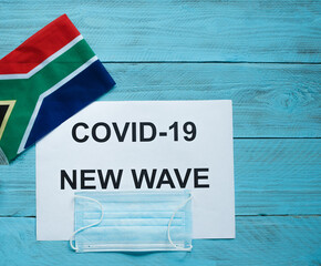Fototapeta na wymiar On a blue wooden background is a sheet with the inscription: covid -19 new wave, a medical mask and the flag of South Africa