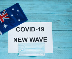 Fototapeta na wymiar On a blue wooden background is a sheet with the inscription: covid -19 new wave, a medical mask and an Australian flag