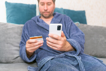 Young man buying fashion items through the Internet on cellphone by using credit card.Online Payment. Frontal portrait of man holding debit card and using smartphone for shopping at home