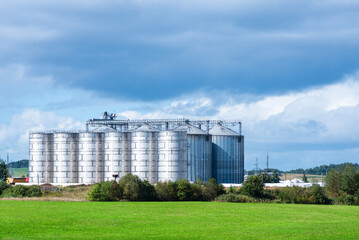 Granary large modern agro-processing plant storage and processing of grain crops. Large metal barrels of grain.Summer cloudy day green field landscape.Copy space.