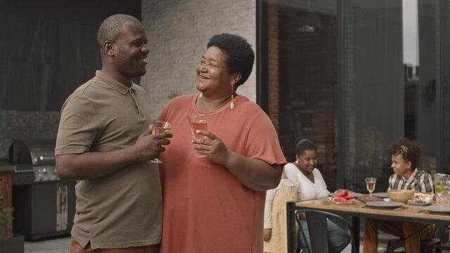 Medium of mature African woman and her adult son drinking wine from glasses, talking, standing on terrace in foreground of family members dining