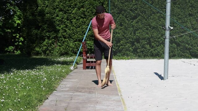 Janitor in a red shirt clears sand from the sidewalk with a broom. A boy in his 20s sweeps the pavement