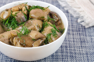 Marinated mushrooms in a white ceramic bowl with dill. The white ethnic fringed napkin on a blue table. Ready-to-eat Suillus. Traditional Ukrainian appetizer of pickled mushrooms
