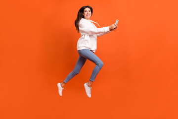 Full length body size view of pretty trendy cheerful girl jumping using gadget chatting isolated over bright orange color background