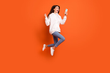 Fototapeta na wymiar Full length body size view of pretty trendy lucky cheerful girl jumping using device win isolated over bright orange color background