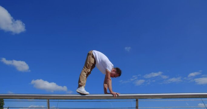 Close up of professional acrobat performing dangerous trick on the railing of the glass bridge against backdrop of city. Caucasian guy walking on hands  on a narrow metal pipe
50 fps slow motion