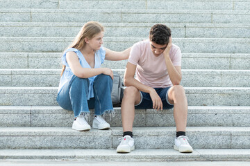 Teenager couple sitting on the stairs. Girlfriend trying to cheer up sad hispanic boyfriend....
