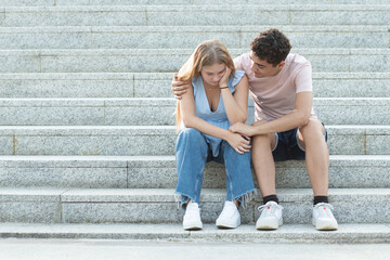 Teenager couple sitting on the stairs. Hispanic boyfriend trying to cheer up and hugging sad...
