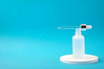 Open frosted transparent glass dropper bottle with serum and a pipette. White podium on blue...