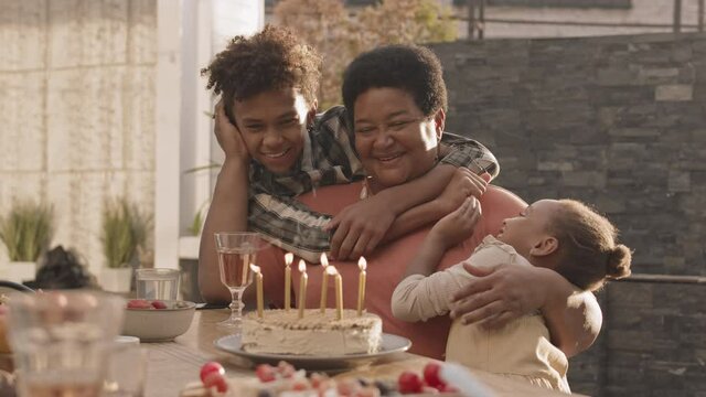 Medium Of Mature African Woman Sitting At Table Outdoors, Her Teenage Male And Five-year-old Female Grandchildren Hugging Her, Then All Blowing Out Candles On Birthday Cake