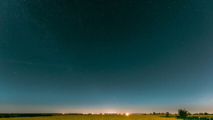 Night Starry Sky Above Summer Field Meadow With Blooming Rapeseed. Glowing Stars And Sunset Sunrise Lights Above Meadow Landscape.