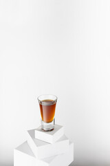 Golden whiskey in shot glass in sunlight with shadow on top of abstract white podiums, alcohol drink in single geometric minimal style, vertical.