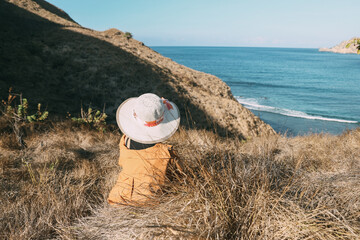 Woman tourist wearing summer hat sitting on savana looking view of the sea and the hills in Padar Island