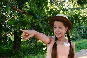 wonderful cheerful young girl in hat stands among green leaves and points her finger at something...