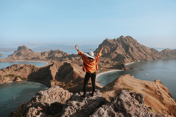 Happy woman traveler in summer hat standing while posing on the top rock mountain with background of hills and seascape