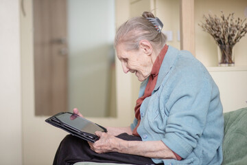 An older woman is talking through a tablet, a modern grandmother, a silver surfer, sitting at home, retirees stayed at home safe during quarantine, online communication on the Internet.
