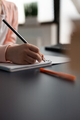 Close up of little girl hand writing on notebook for online math class learning at home desk. Student holding pencil to write exercise on copybook for educational lecture knowledge