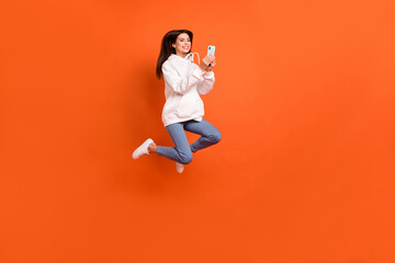 Full length body size view of pretty trendy cheerful girl jumping using device 5g isolated over bright orange color background
