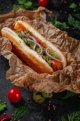 Delicious juicy hot dog with cheese sauce, tomato and fresh herbs in assortment, sandwich on the menu of a fast food restaurant on a dark stone table. Healthy option of fast food.