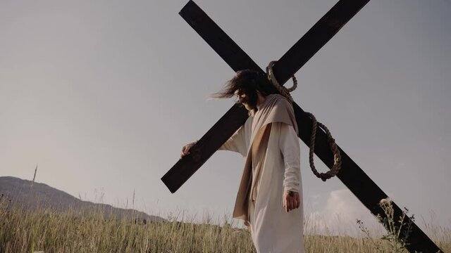 Jesus carries the cross on his back. He walks on the grass, against the background of the hills. The wind is blowing, the sun is shining.