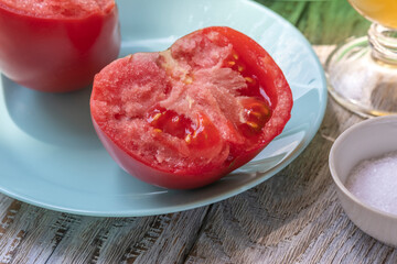 Large ripe, fleshy tomato, broken in half by hands. Texture of pulp is clearly visible. Half tomato is lying on turquoise plate. Salt shaker with salt is visible. On wooden table