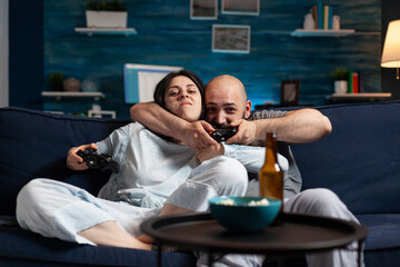 Determinated enthusiastic couple winning video games, hugged each other sitting on sofa playing, excited scream enjoying online soccer competition. Competitive happy gamers using gaming joysticks