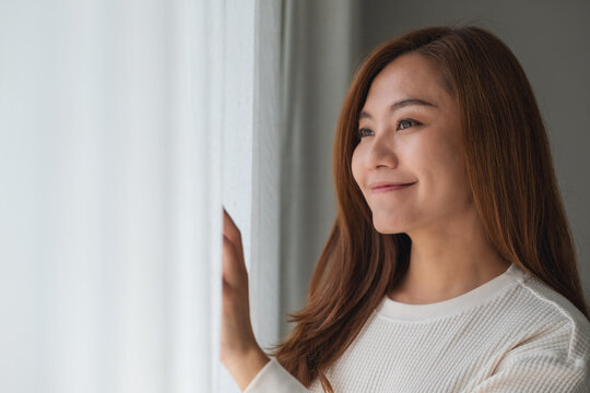 A beautiful young asian woman touching curtain and looking outside the window