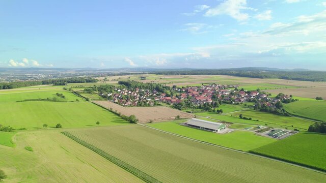 Aerial view of a metall cow shed next to a beautiful German village surrounded by green meadows and summer landscape. Tilting and panning drone shot.