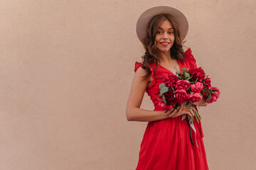 Medium shot portrait of magnificent girl with cherry lipstick in light summer dress and pink peonies bouquet looks happy. Curly long brunette hair under the hat. Solid background behind the woman 