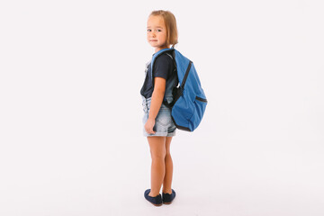 Little girl with blond hair dressed in a denim overalls and a blue t-shirt, with a backpack ready for going back to school, on her side, on white background