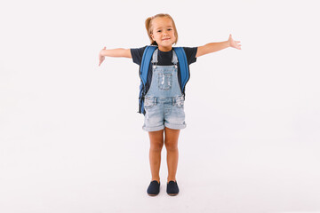 Little girl with blond hair dressed in a denim overalls and a blue shirt, with a backpack ready for going back to school, with her arms open, very happy, on white background